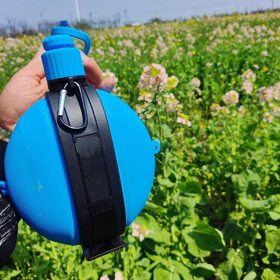 A hand holds a bright blue collapsible silicone camping kettle outdoors in a field of green plants.