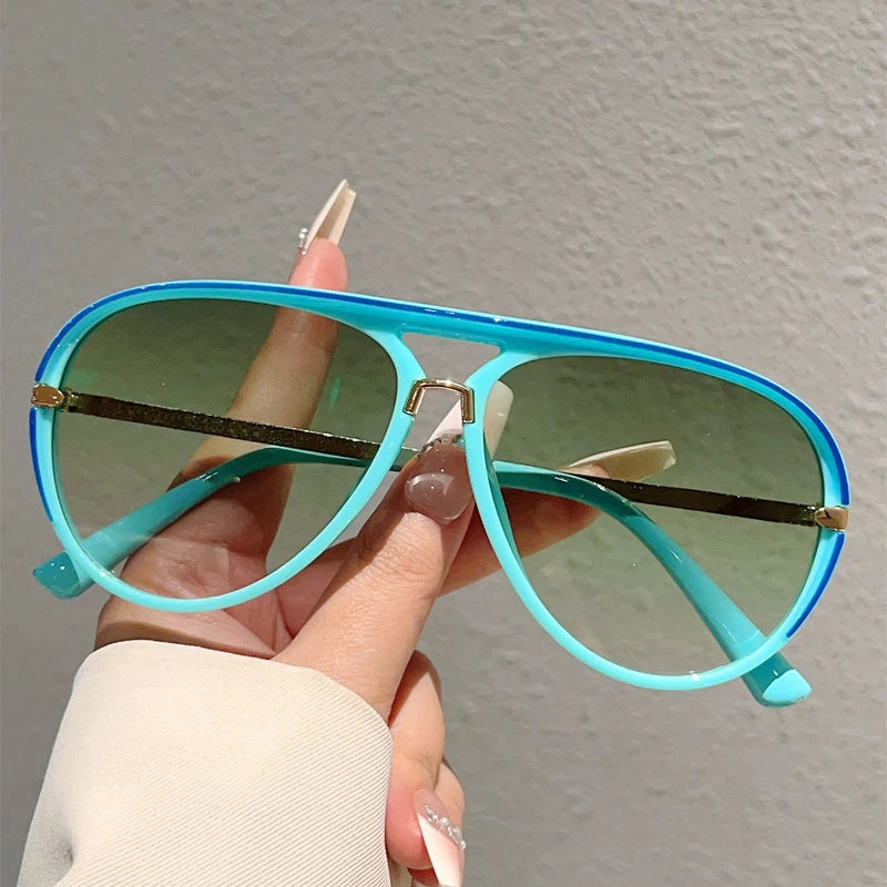 A hand holds a pair of light blue gradient sunglasses with gold accents against a plain gray background.