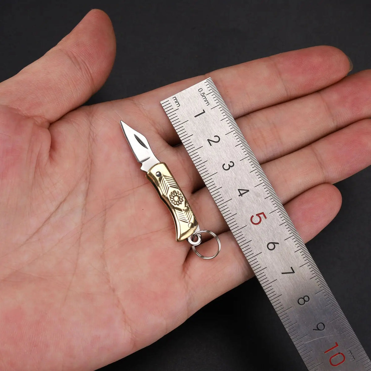 Un mini cuchillo plegable de latón para llavero está apoyado en una regla en la mano de una persona.