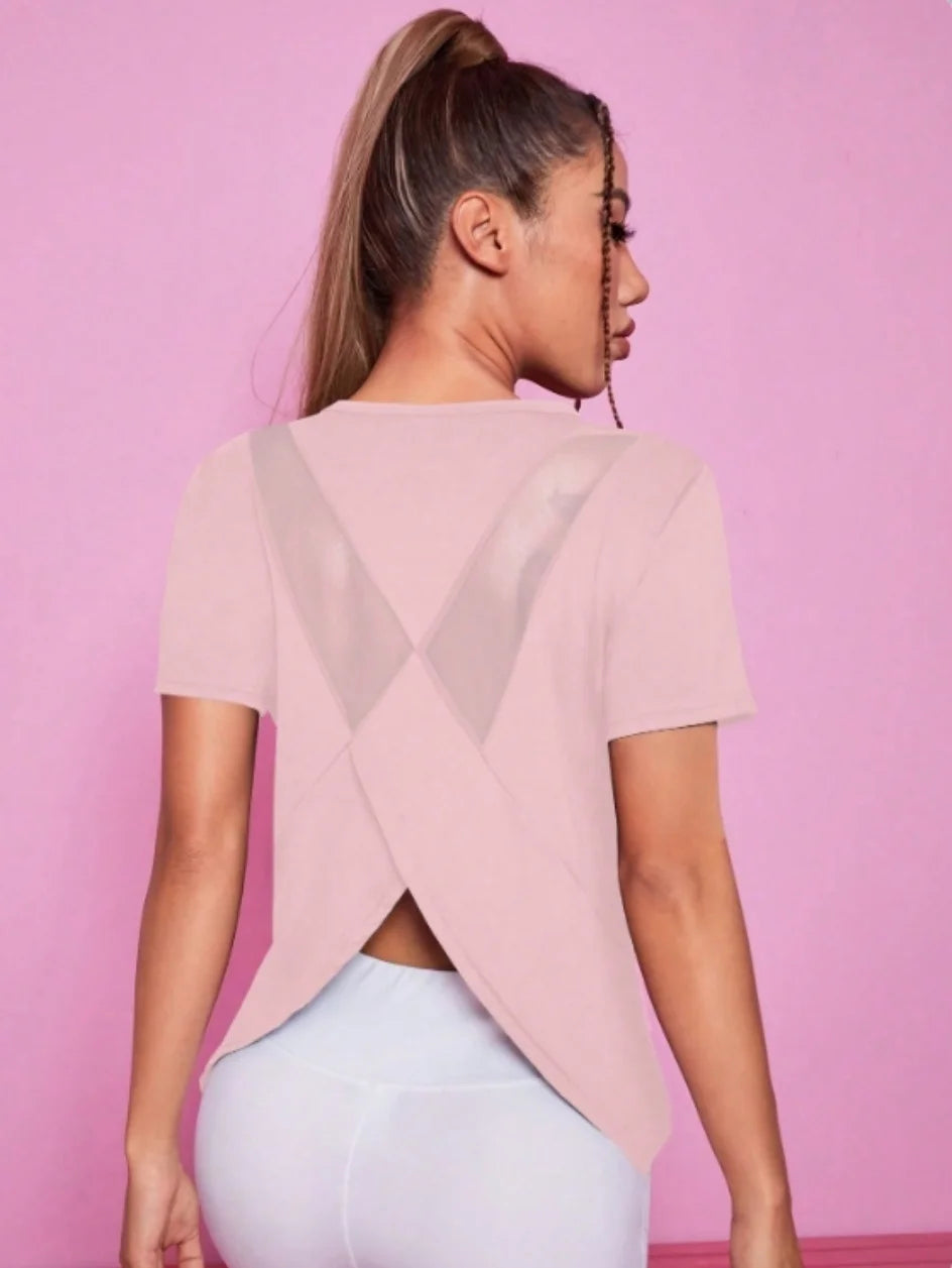 A woman wearing a light pink breathable summer yoga and fitness t-shirt with an X-shaped cutout against a pink background.