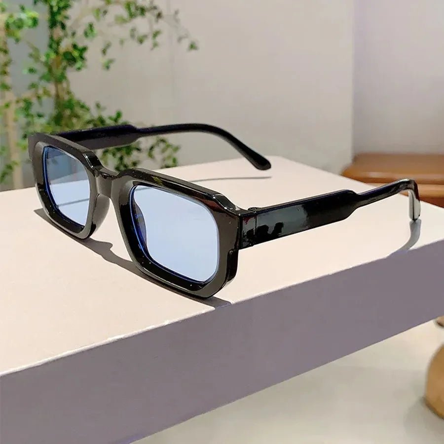 Lunettes de soleil à monture rectangulaire noire rétro posées sur une surface blanche.