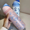 A hand holds a pink water bottle with blue floral designs and a light blue lid against a white background.