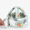 A hand holds a soft, adjustable safety helmet for toddlers with a light green fabric featuring various animal and geometric p