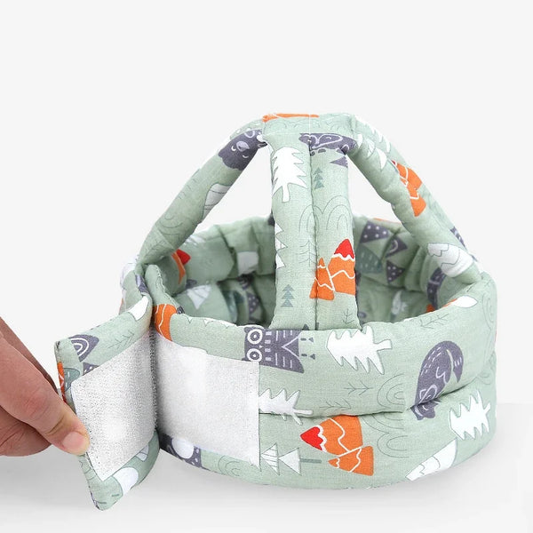 A hand holds a soft, adjustable safety helmet for toddlers with a light green fabric featuring various animal and geometric p