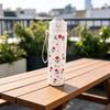 A white tumbler decorated with red strawberry and cherry flowers sits on a wooden table outdoors.