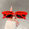 Une main tient des lunettes de soleil rectangulaires rétro rouges vif sur un fond clair.