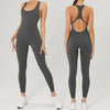 A woman models a dark gray backless fitness jumpsuit in a full-length shot.