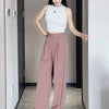 A woman models loose pink yoga pants and a white sleeveless top in a hallway.