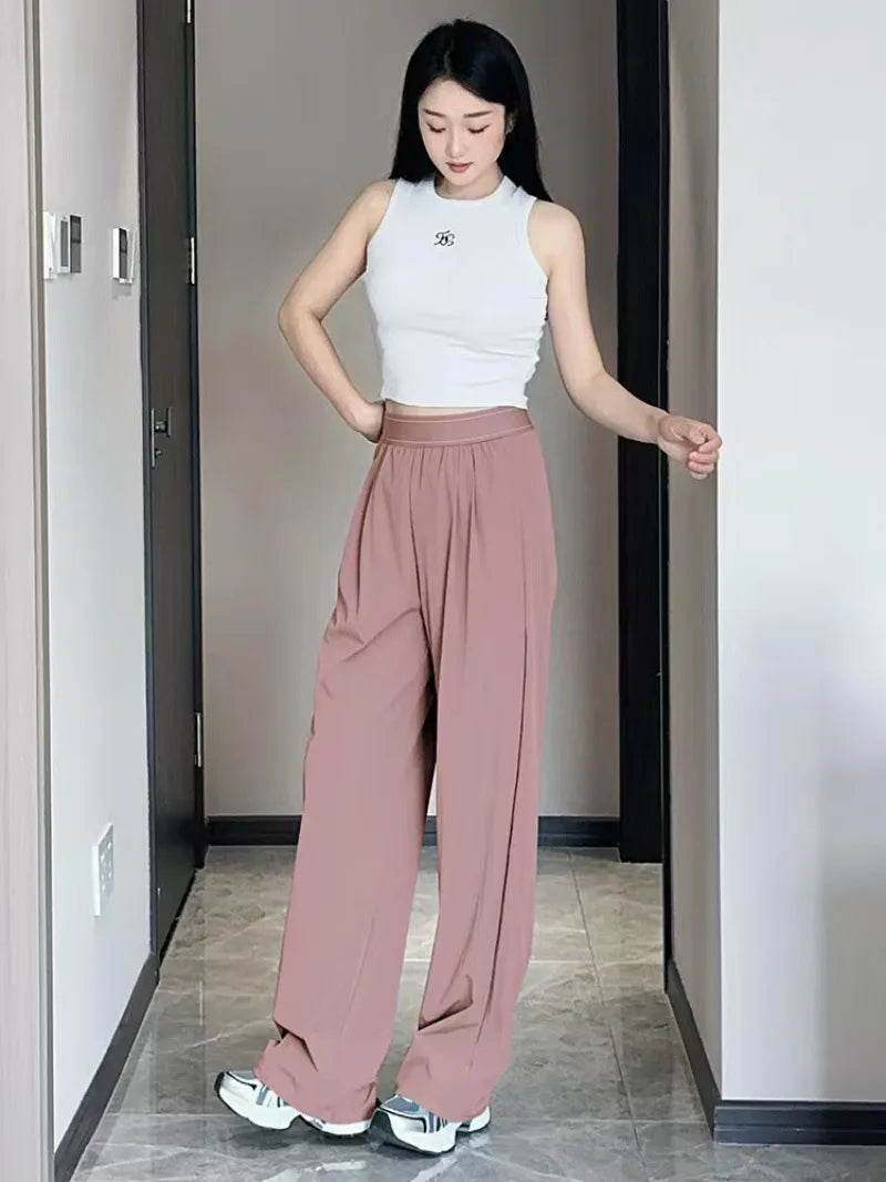 A woman models loose pink yoga pants and a white sleeveless top in a hallway.