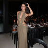 A model wearing an elegant, olive green maxi dress with a draped cut-out back, posing in a dimly lit restaurant setting.