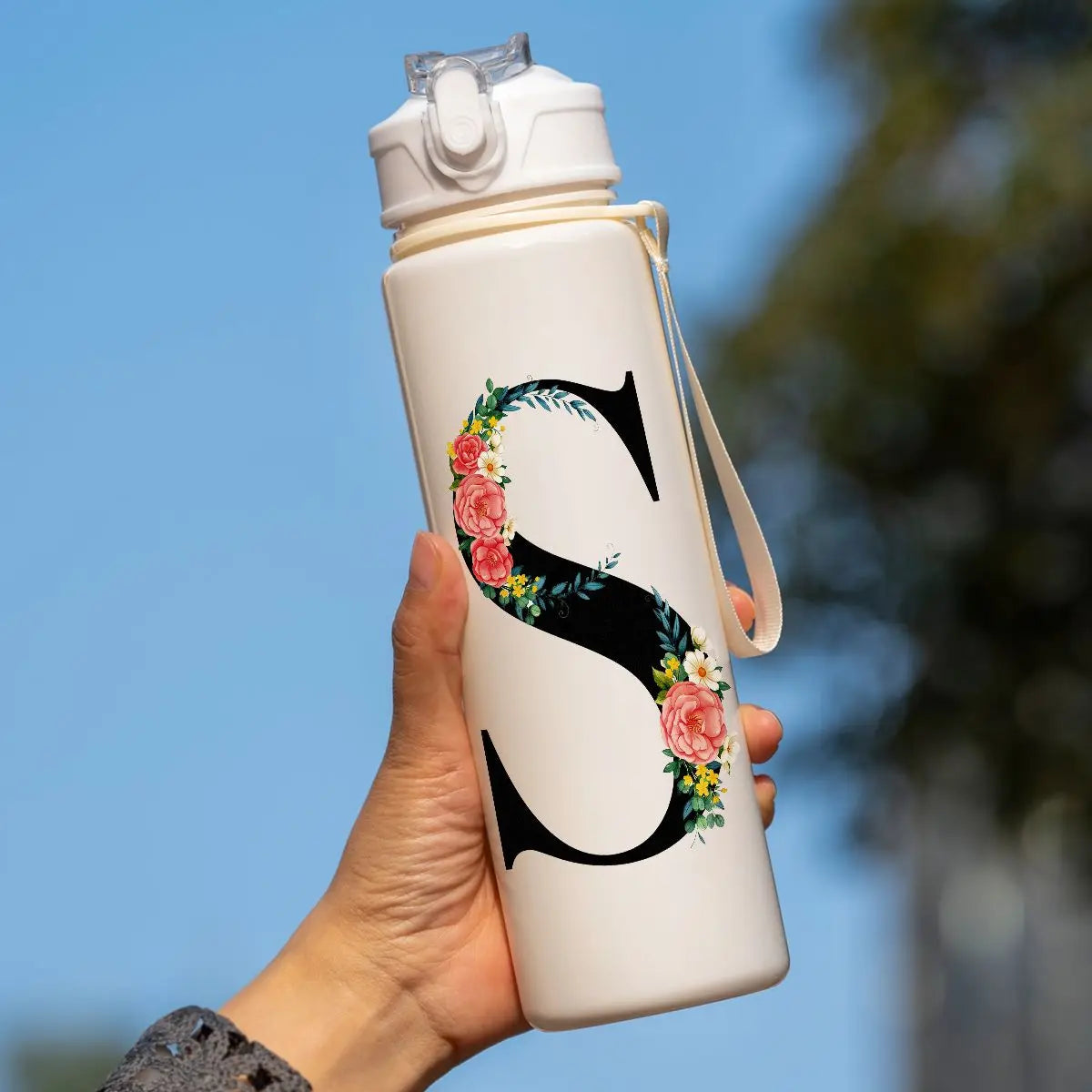 A hand holds a white insulated straw tumbler with a large black initial S decorated with pink and yellow flowers against a bl