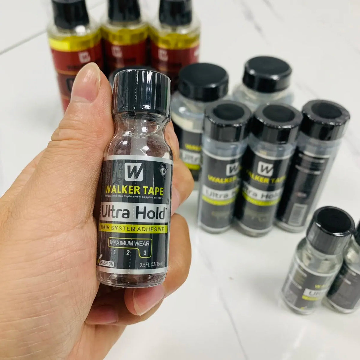 A hand holds a bottle of Walker Tape Ultra Hold Hair System Adhesive for lace wigs and hair replacement.