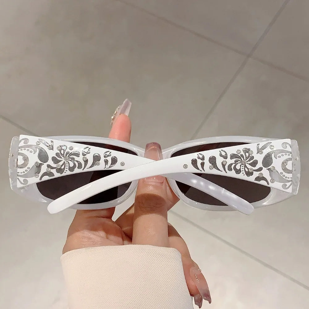 A hand holds a pair of white sunglasses with silver rhinestone detailing against a light background.