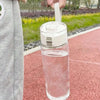 A hand is holding a large capacity transparent sports water bottle with a white cap outdoors.