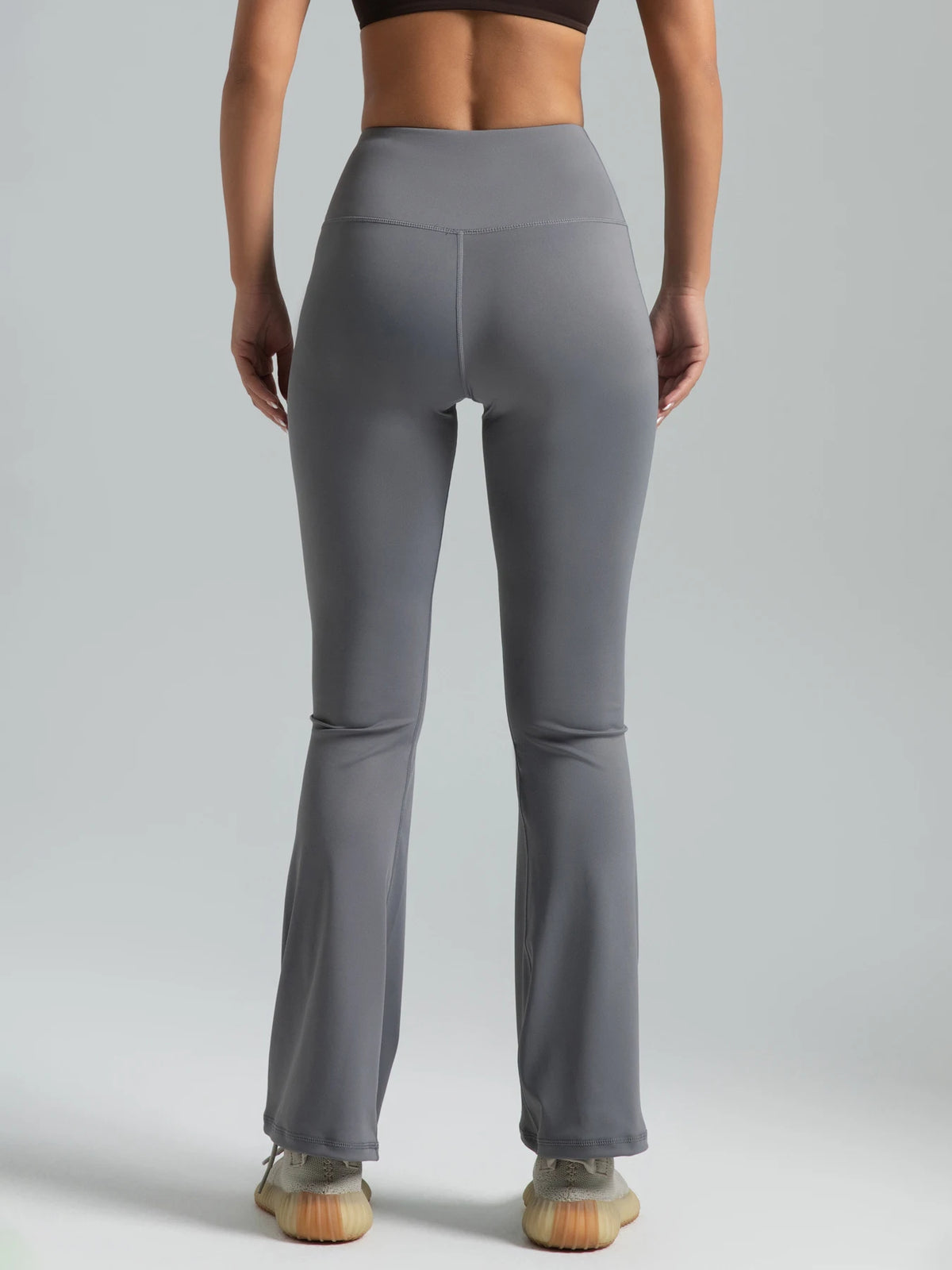 A person wearing high-waisted grey bootcut yoga pants and a black sports bra is standing against a light gray background.