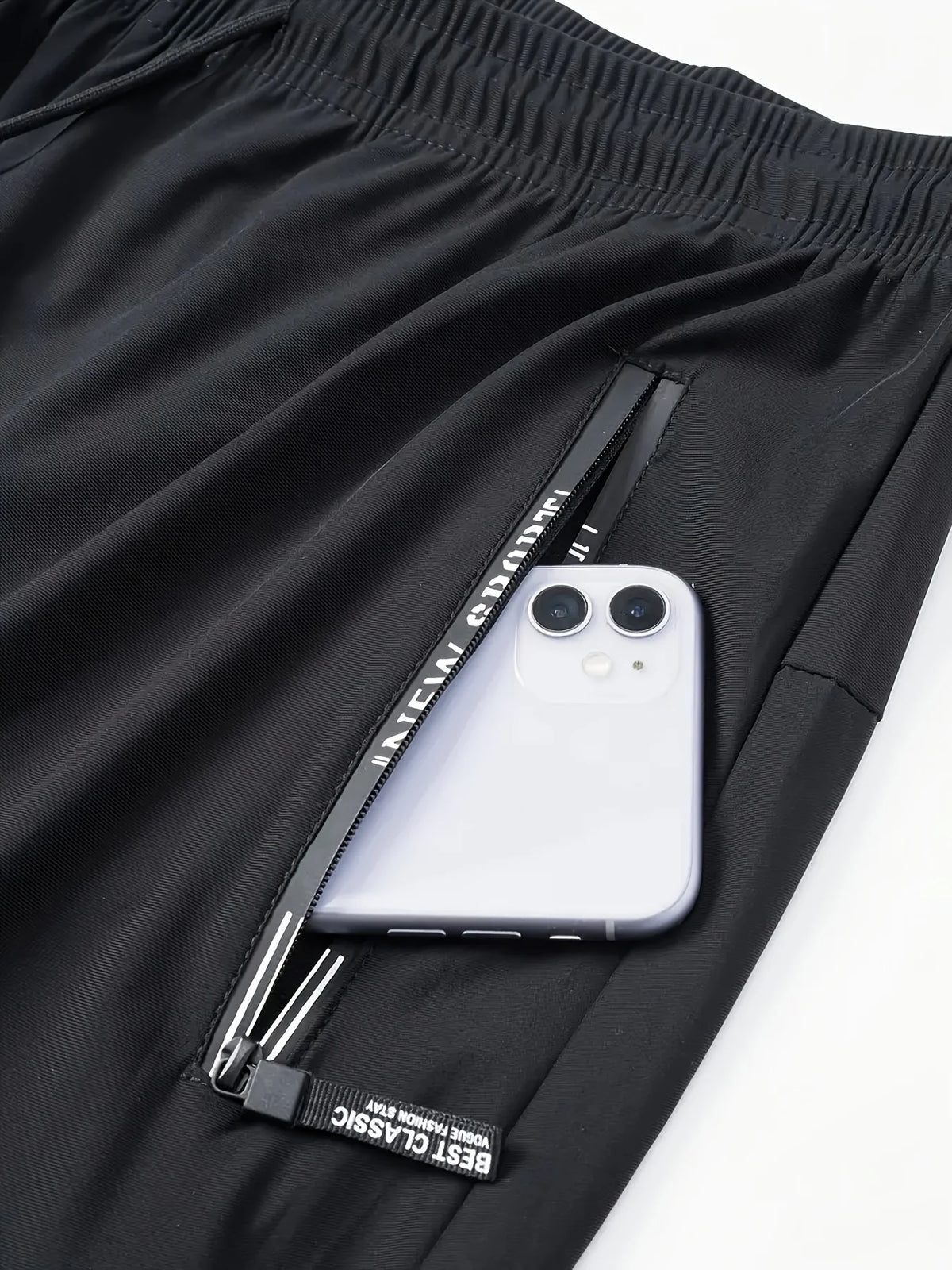 A white smartphone is placed inside a zippered pocket of black breathable men's casual jogger pants.