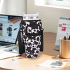 A black adjustable pouch carrier for a water bottle is shown on a wooden desk next to a laptop and papers.