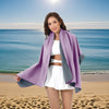 A young woman wearing a purple microfiber quick-dry beach towel and white outfit stands on a sandy beach with the ocean in th
