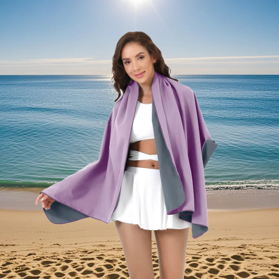 A young woman wearing a purple microfiber quick-dry beach towel and white outfit stands on a sandy beach with the ocean in th