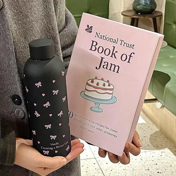 A pink booklet titled National Trust Book of Jam is held by a person next to a black insulated travel mug.