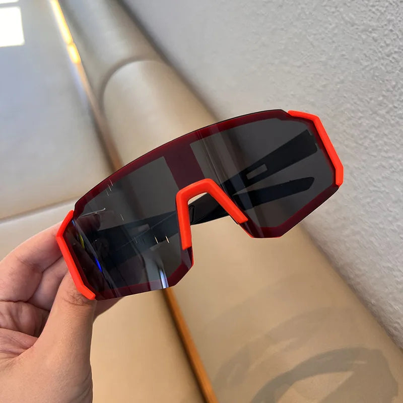 A hand holds a pair of orange-framed cycling sunglasses with dark lenses against a light background.