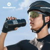 A man wearing a black helmet and sunglasses holds a black Rockbros sports water bottle against a bright sky.
