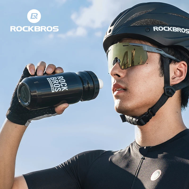 A man wearing a black helmet and sunglasses holds a black Rockbros sports water bottle against a bright sky.