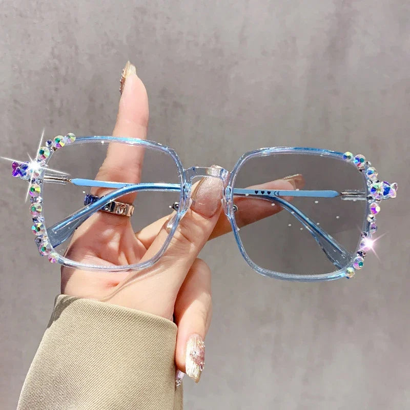 A hand holds a pair of light blue photochromic diamond style eyeglasses with sparkling accents against a gray background.