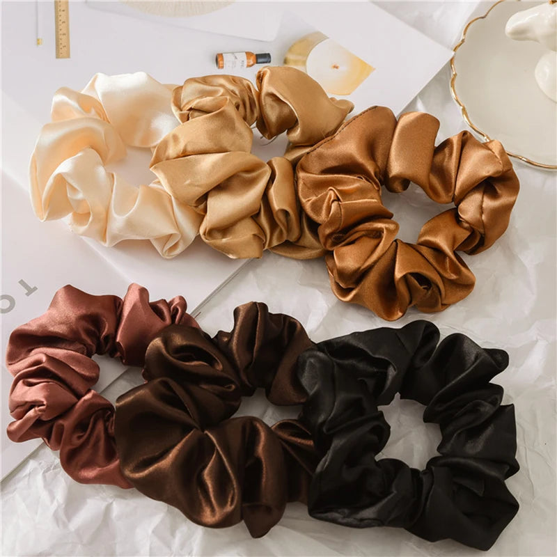 A collection of satin scrunchies in various shades including beige, gold, bronze, and black laid out on white paper.
