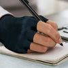 A hand wearing a black compression fingerless glove is writing in a notebook.