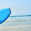 A man wearing a blue shirt holds a bright blue surfboard near the ocean water.