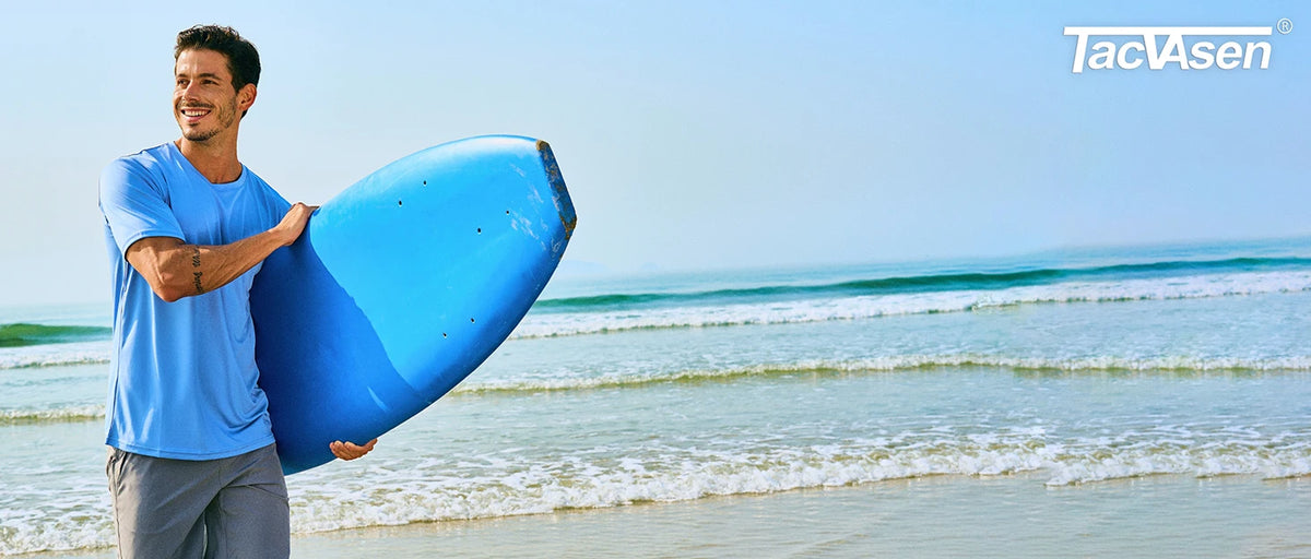 A man wearing a blue shirt holds a bright blue surfboard near the ocean water.