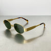 Gold-toned metal oval sunglasses with olive green lenses resting on a gray surface.