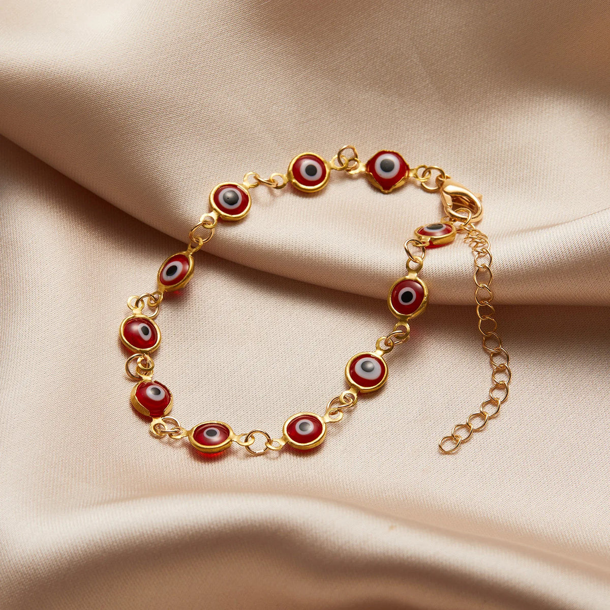 Gold charm bracelet featuring round evil eye beads with red and white designs on a soft beige fabric.