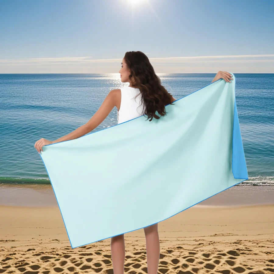 A woman holds up a light blue microfiber quick-dry beach towel against the ocean backdrop.