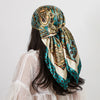 A woman wearing a teal and gold paisley silk shawl scarf tied around her neck, visible over a sheer white top.