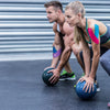 Two muscular individuals performing a physical exercise with a blue medicine ball in a gym setting.