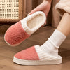 A pair of pink and cream plush fleece slippers being held by a hand, shown on a wooden floor.