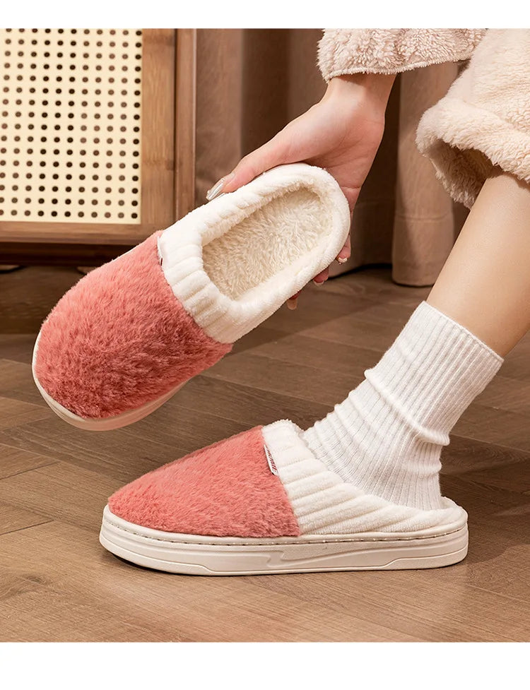 A pair of pink and cream plush fleece slippers being held by a hand, shown on a wooden floor.
