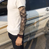 A person wearing a white t-shirt and dark jeans, displaying a full sleeve tattoo on their arm next to a silver car.