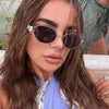 A woman with long brown hair wearing classic oval metal sunglasses and a light blue top.