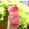A hand holds a pink leak-proof shaker bottle filled with an orange liquid against a bright green, leafy background.