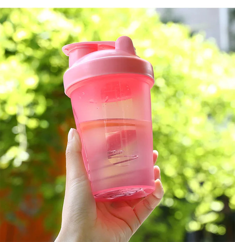A hand holds a pink leak-proof shaker bottle filled with an orange liquid against a bright green, leafy background.