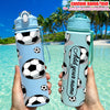 A light blue personalized football water bottle with a black and white soccer ball pattern is held against a tropical beach b