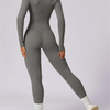 Model trägt einen figurbetonten, langärmeligen grauen Yoga-Jumpsuit mit hohem Kragen und Langhosen.