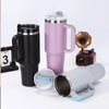 Tasse isotherme en acier inoxydable rose avec anse et une tasse noire, à côté d'une petite caméra.