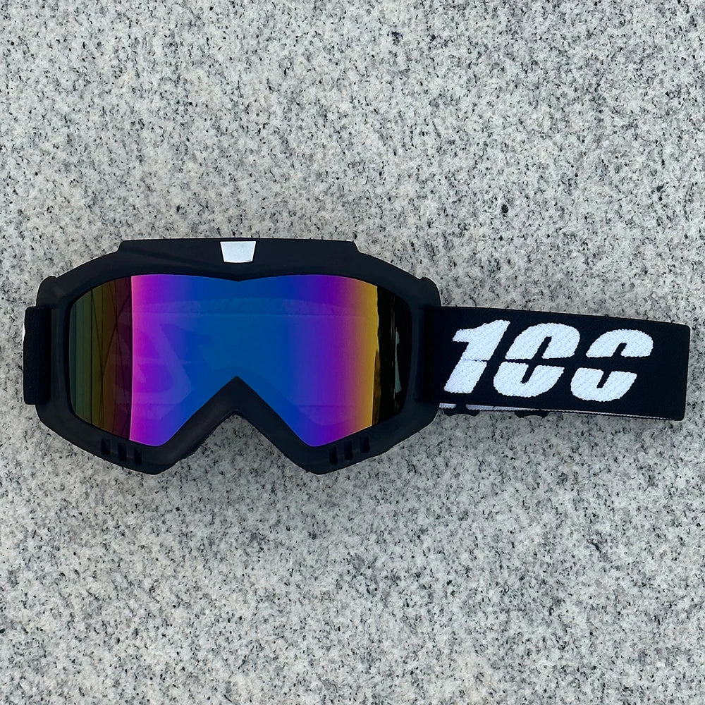 A pair of black motorcycle goggles with a reflective purple and blue lens resting on a gray surface.