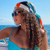 A woman with curly hair wearing an elegant satin square scarf tied around her head outdoors near the sea.