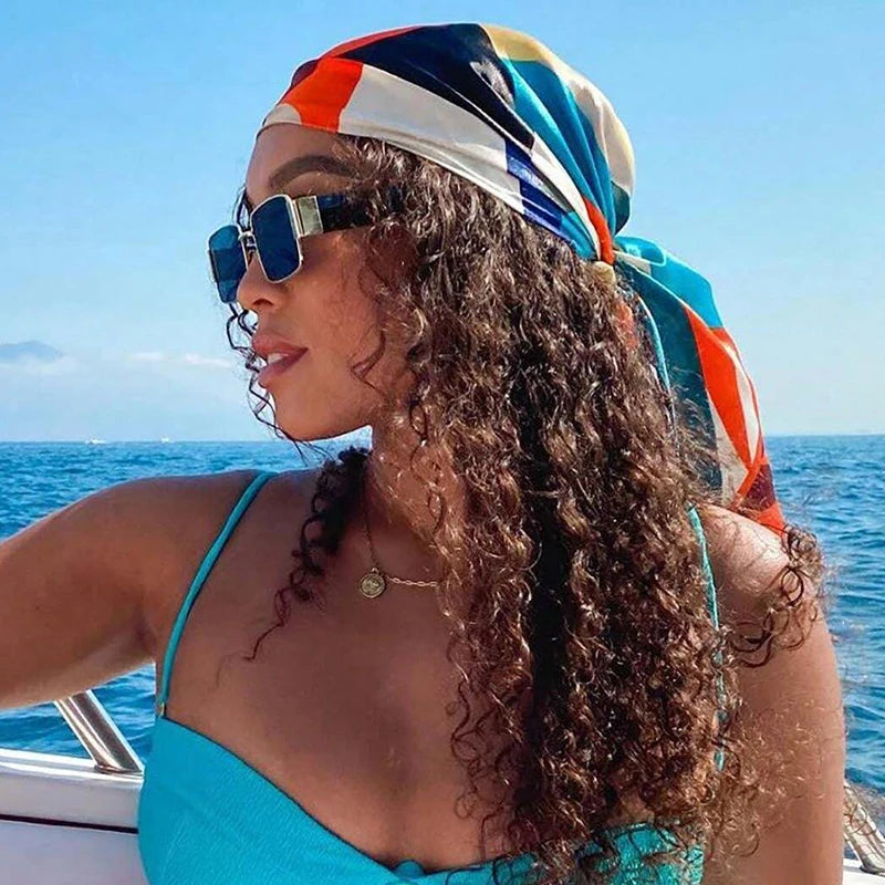 A woman with curly hair wearing an elegant satin square scarf tied around her head outdoors near the sea.