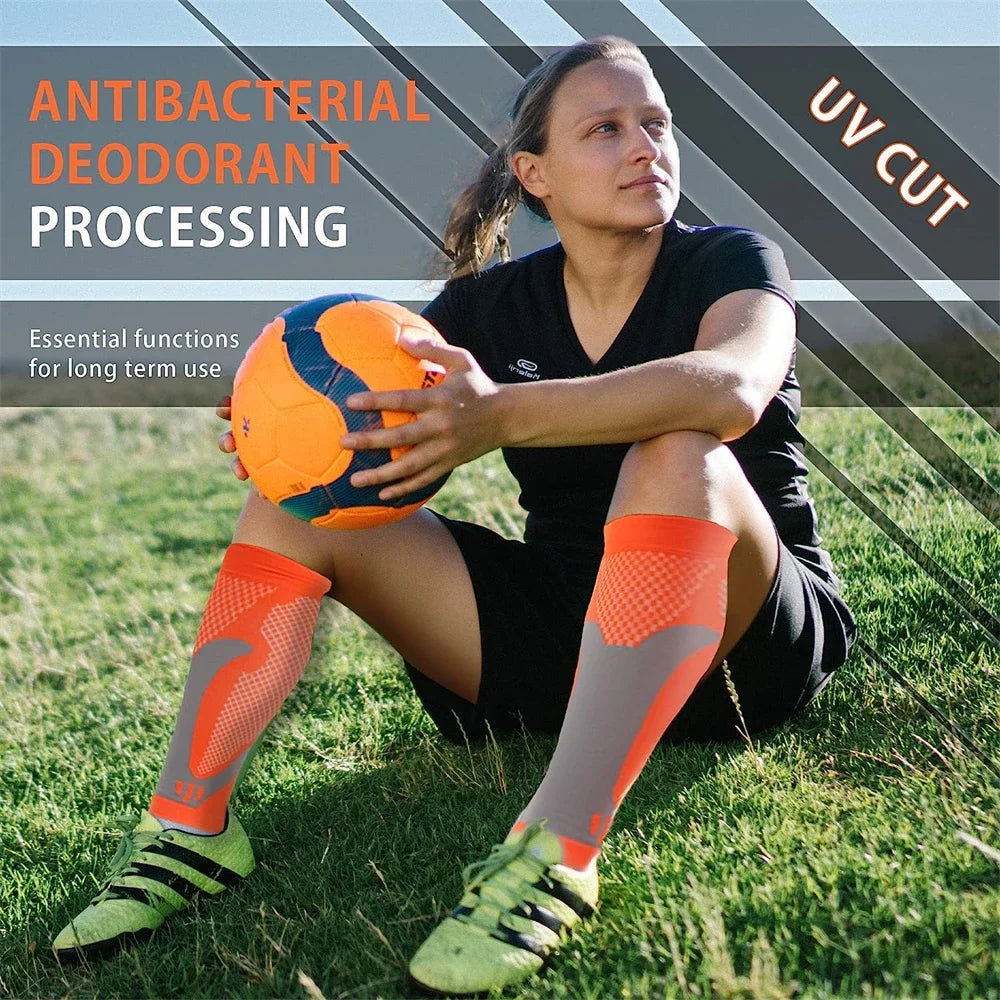 A woman wearing orange calf compression sleeves is sitting on grass with an orange ball.